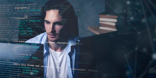A man looking at one of the two computer monitors in front of him with a few books behind him